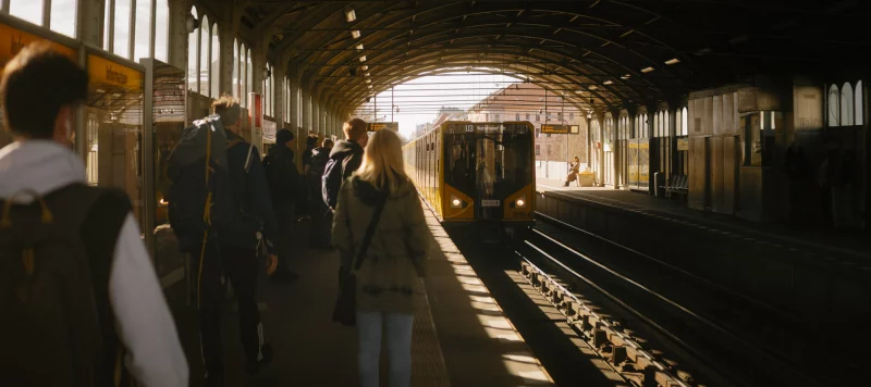 Wartende Fahrgäste auf dem Bahnsteig des Hochbahnhofs Prinzenstraße in Berlin bei warmem Sonnenlicht. Ein gelber U3-Zug der Berliner U-Bahn fährt in den historischen Stahlbogenbahnhof ein. Links ist ein Informationsschild zu sehen.
