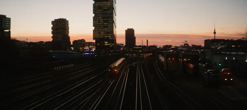 Blick von einer Brücke auf die Gleisanlagen am U-Bahnhof Warschauer Straße in Berlin bei Sonnenuntergang. Im Vordergrund verlaufen mehrere Bahngleise, auf denen ein beleuchteter U-Bahn-Zug zu sehen ist. Im Hintergrund die Berliner Skyline mit modernen Hochhäusern, dem Berliner Fernsehturm und einem rosa-orangefarbenen Abendhimmel.