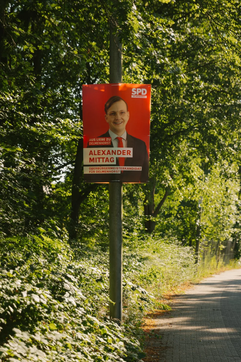 Es ist das Wahlplakat des SPD-Oberbürgermeister-Kandidaten für Delmenhorst Alexander Mittag zu sehen. Es hängt an einem Laternenmast an einer Straße. Im Hintergrund sind die Blätter vieler Bäume zu sehen.