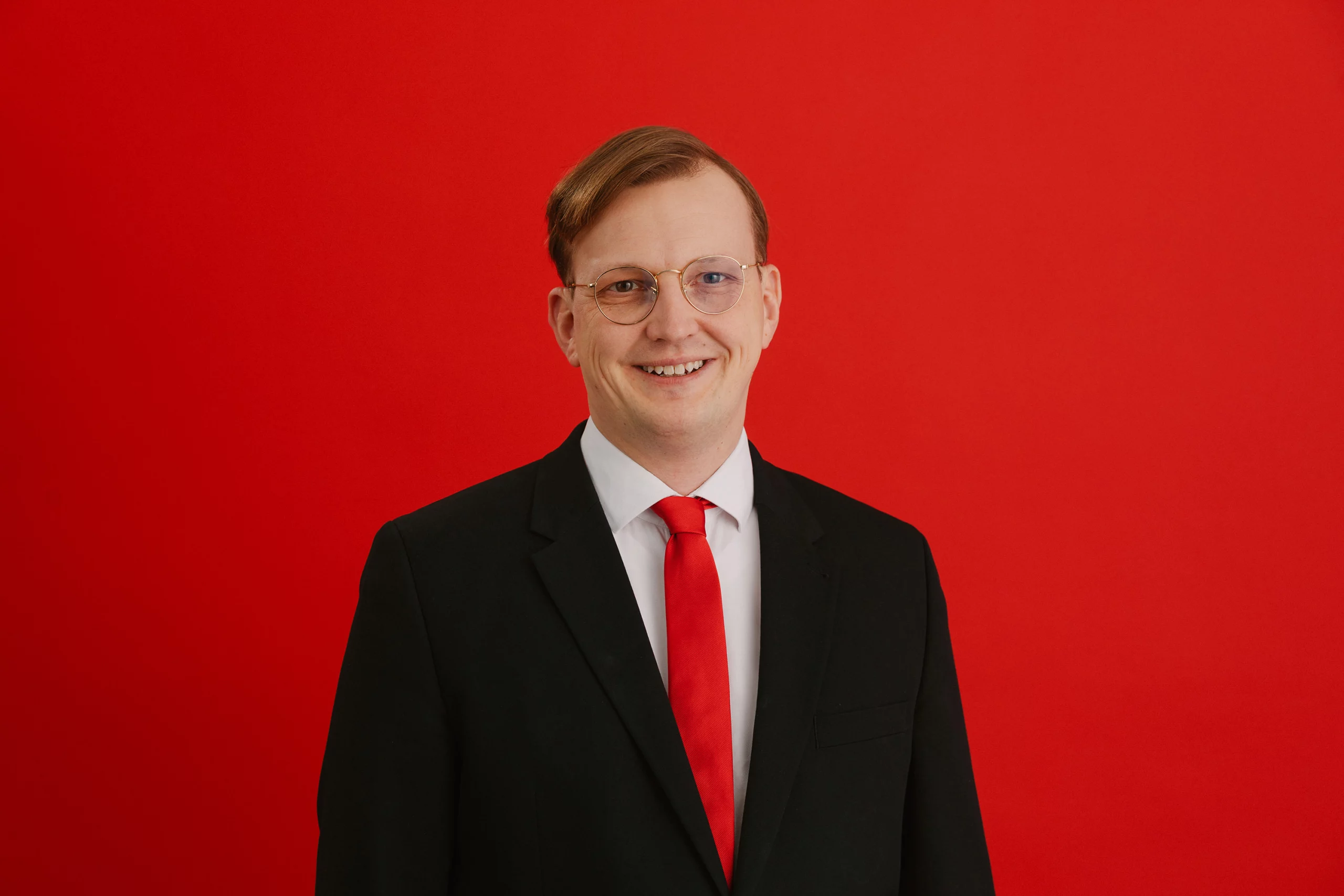 Portraitfoto von Alexander Mittag, dem Oberbürgermeisterkandidaten der SPD für Delmenhorst bei der niedersächsischen Kommunalwahl im September 2026. Er trägt einen schwarzen Anzug, ein weißes Hemd und eine Krawatte in SPD-rot. Der Hintergrund ist ebenfalls im gleichen Feuerrot. Die Haare sind zu einem Seitenscheitel gekämmt und er lächelt freundlich in die Kamera.